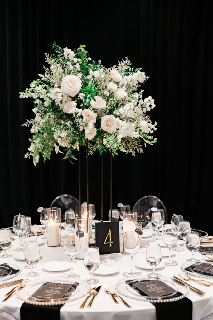 Tall reception centerpiece of white floral and greenery on brass stand.