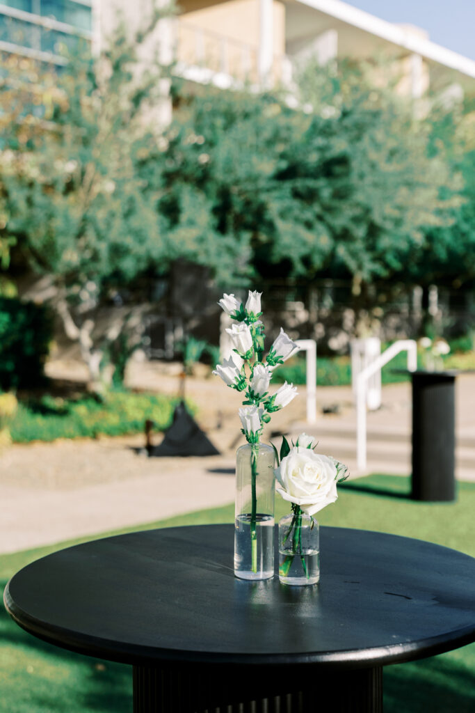 Two modern clear glass bud vases with white flowers in them on a black high top table.