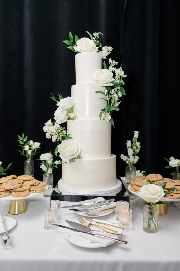 Four tiered white wedding cake with white flowers added, bud vases, votive candles, and cookie trays on table around cake.
