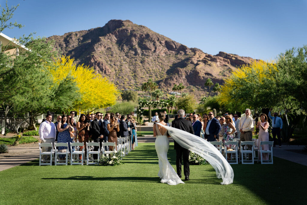 Viewing back of bride entering outdoor wedding ceremony aisle with man walking towards groom at front altar space with Mountain Shadows in the background.
