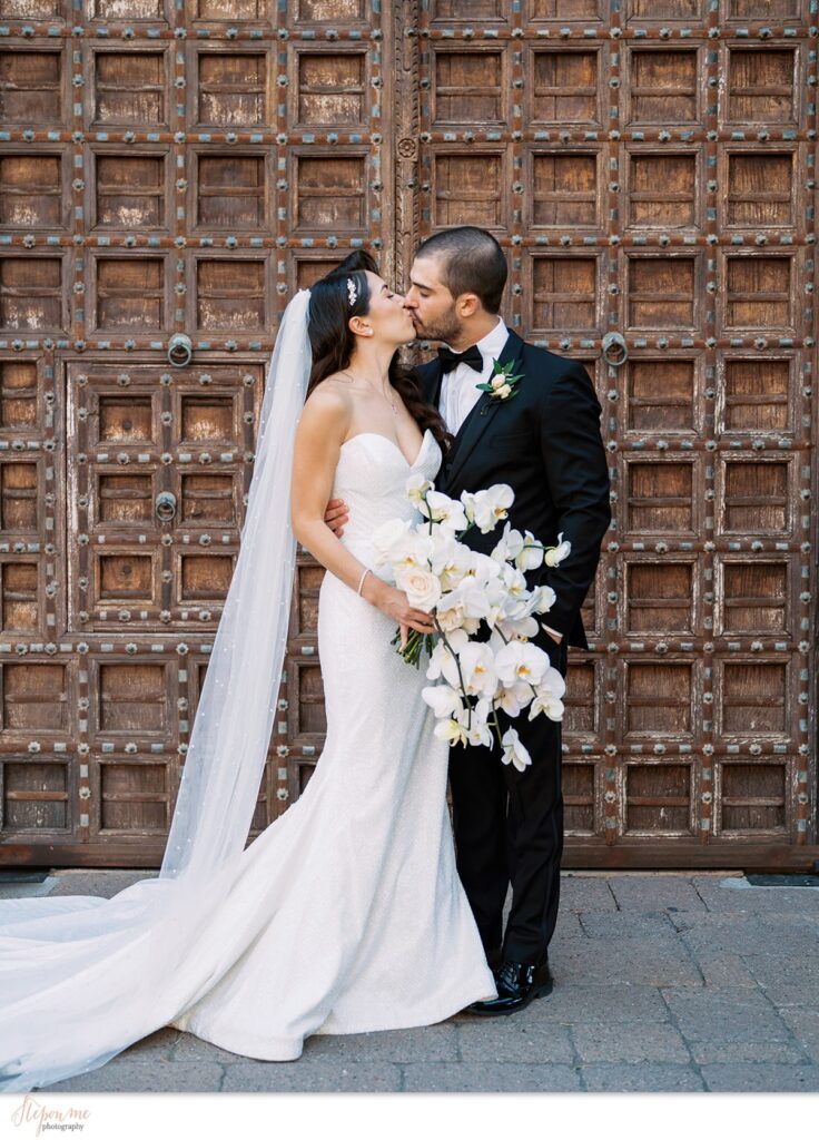 Bride and groom kissing in front of large wooden doors, bride holding lush bouquet of white roses and orchids.