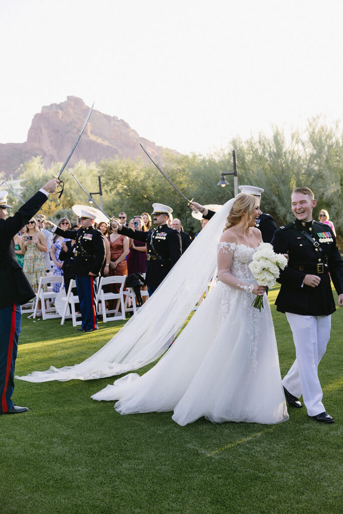 Bride and groom in military uniform with her arm in his, smiling, bride holding bouquet of roses with long stems walking under military sword tunnel at outdoor wedding ceremony.