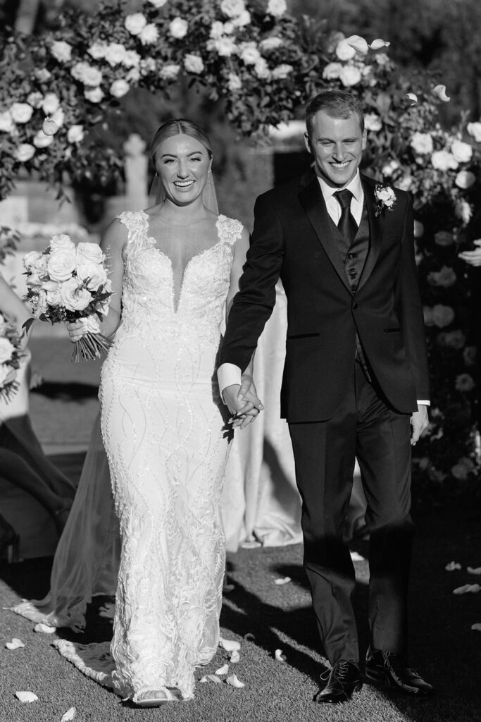 Bride and groom holding hands, smiling, walking down wedding ceremony aisle with floral and greenery arch behind them.