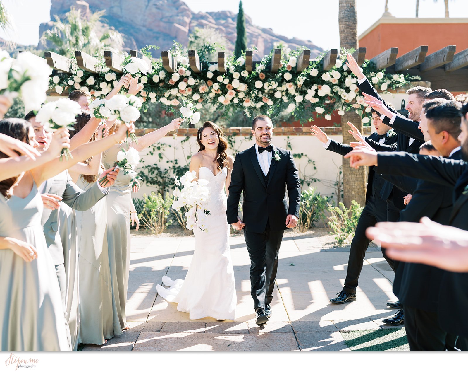 Bride and groom holding hands, smiling, walking between groomsmen and bridesmaids holding hands up above in celebration.