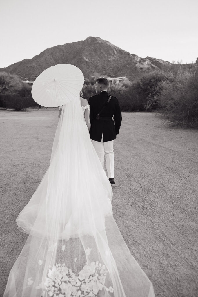 Bride and groom walking away in desert landscape, bride with long sheer train and holding white open umbrella.