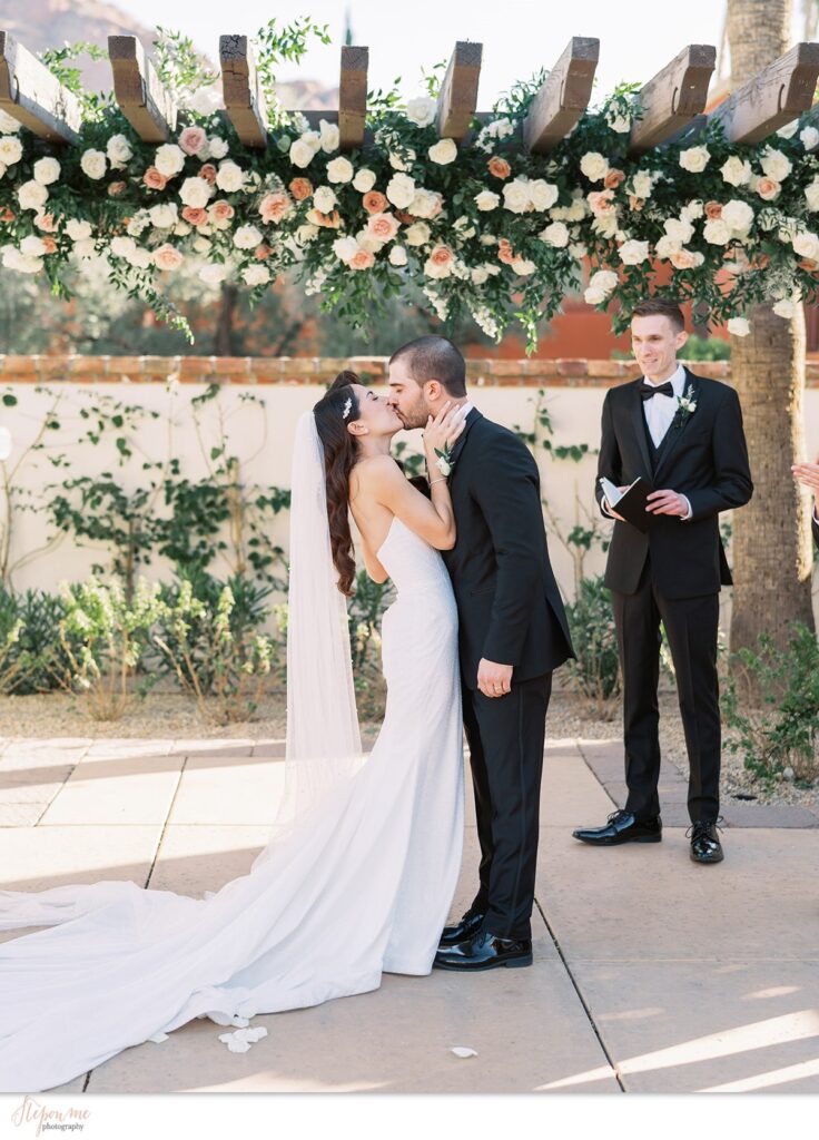 Bride and groom kissing under Omni resort pergola with floral installation of white and pink, mauve flowers.