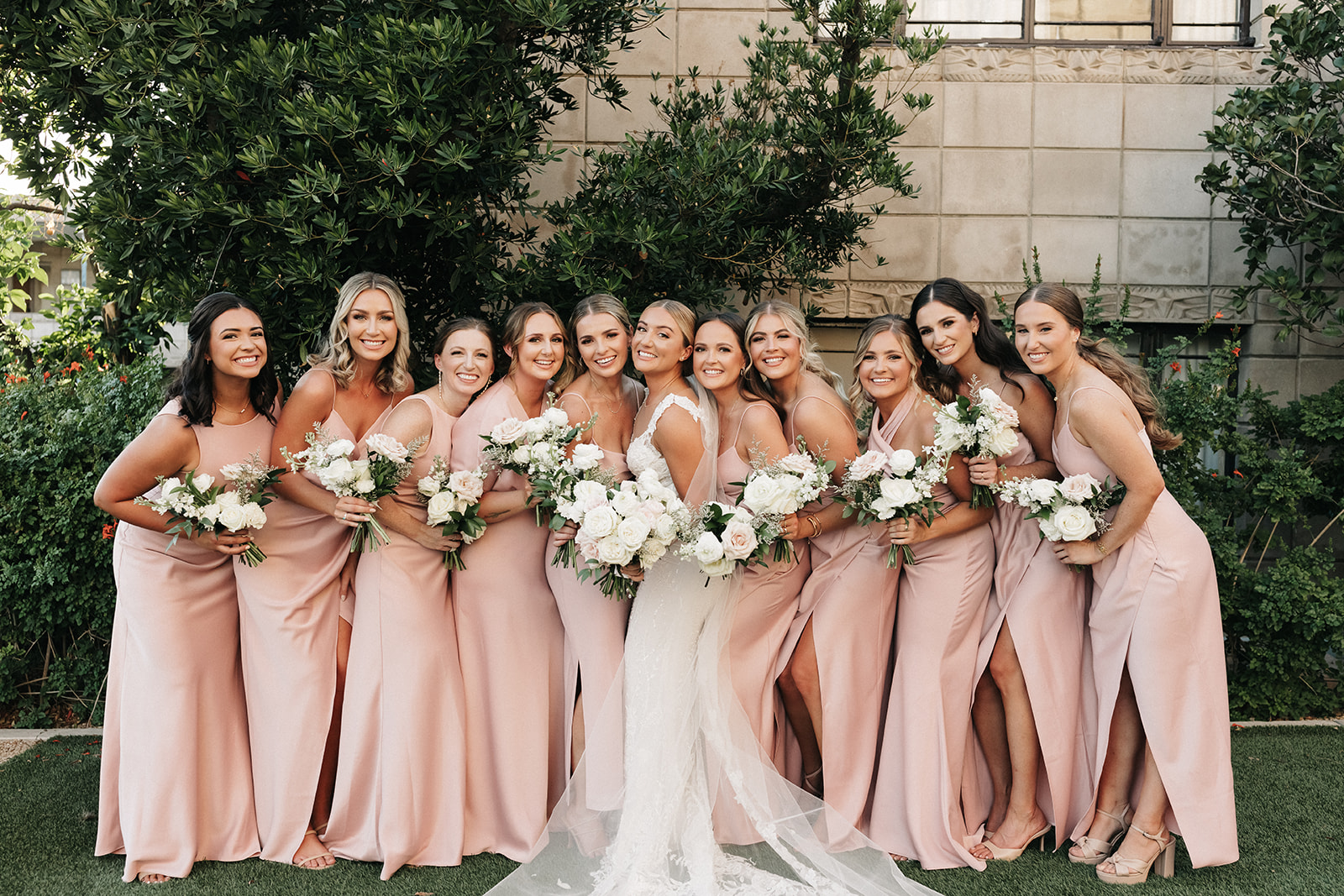 Bride in center of line of bridesmaids wearing blush gowns, all smiling, holding soft colors bouquet flowers.