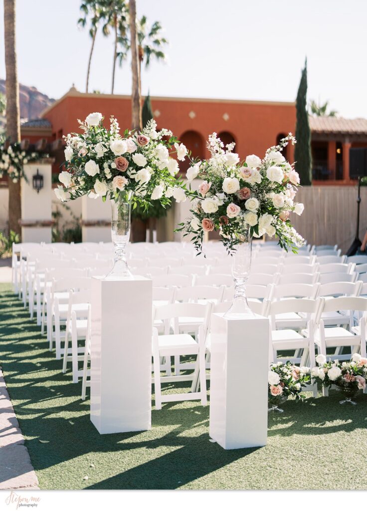 Outdoor wedding ceremony with two tall floral arrangements in glass vases resting on white acrylic pillars.