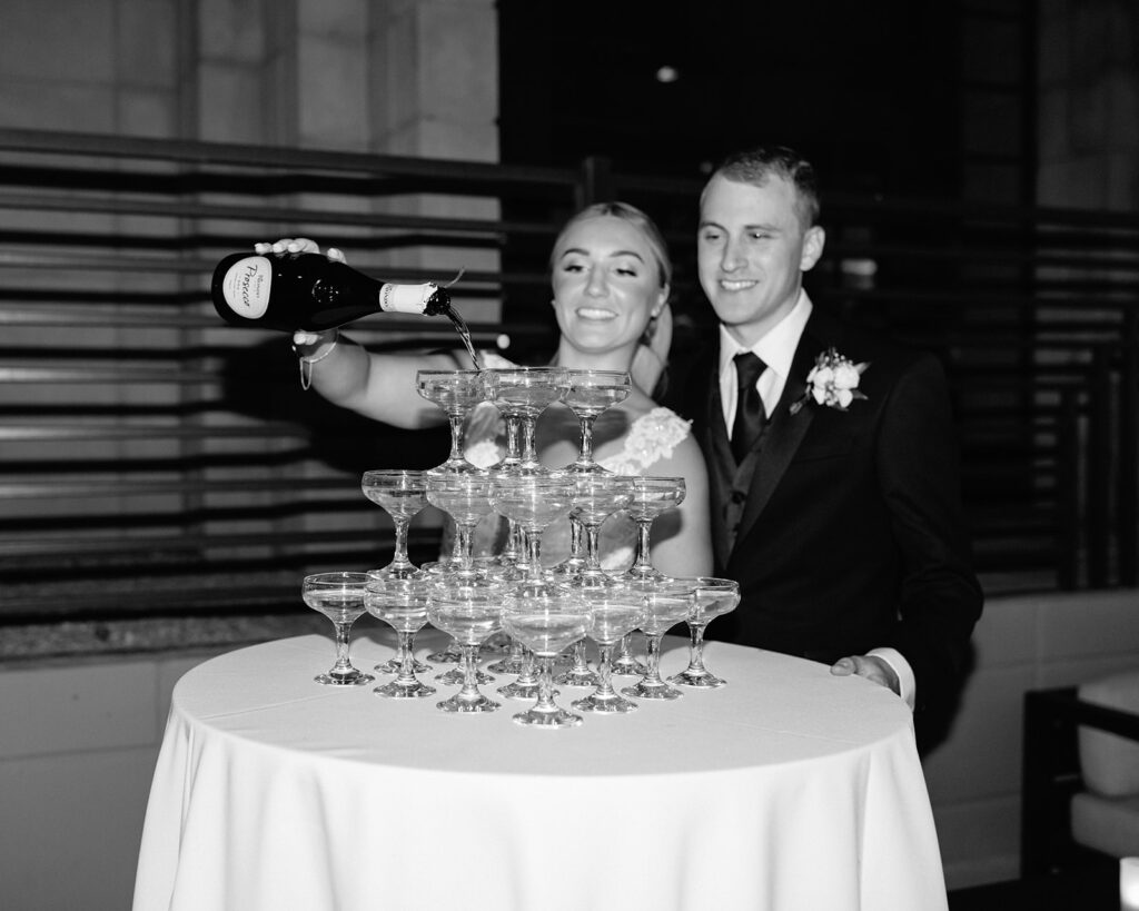 Bride pouring champagne over champagne glasses tower on table with groom standing behind her.