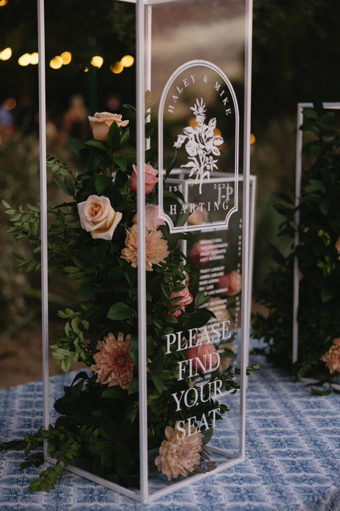 Custom wedding escort sign clear column with floral and greens in center.