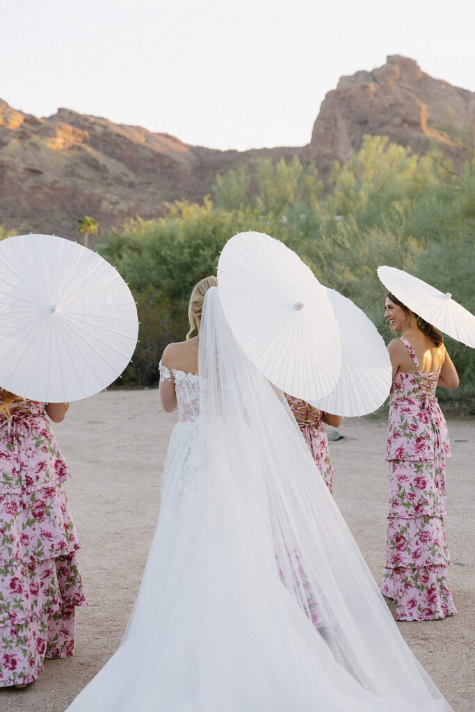 Bride and bridesmaids walking in desert landscape holding open paper umbrellas over them.