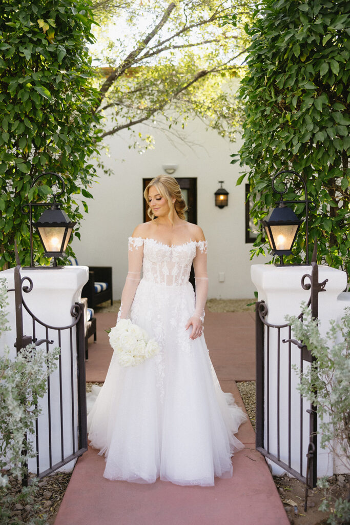 Bride holding white roses bouquet to her side standing in short wall entryway at El Chorro, smiling and looking down.