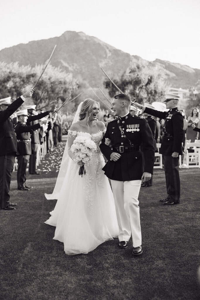 Bride and groom in military uniform with her arm in his, smiling, bride holding bouquet of roses with long stems walking under military sword tunnel at outdoor wedding ceremony.