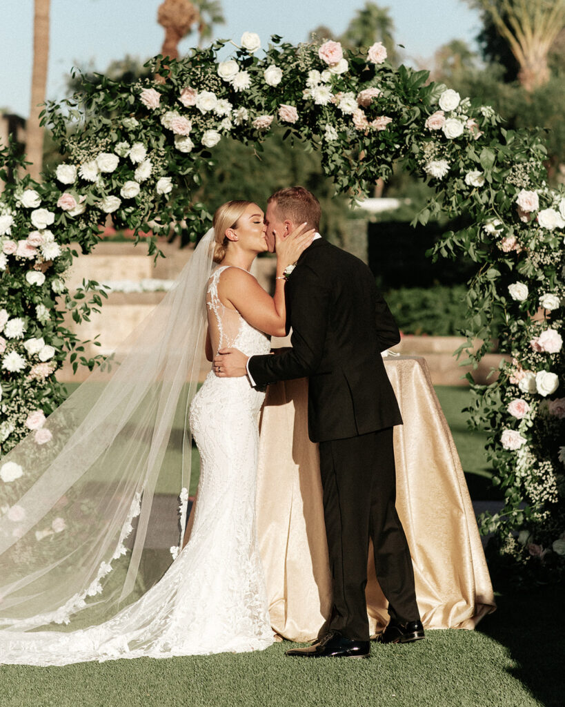 Bride and groom kissing under wedding ceremony arch of greenery and flowers outside.