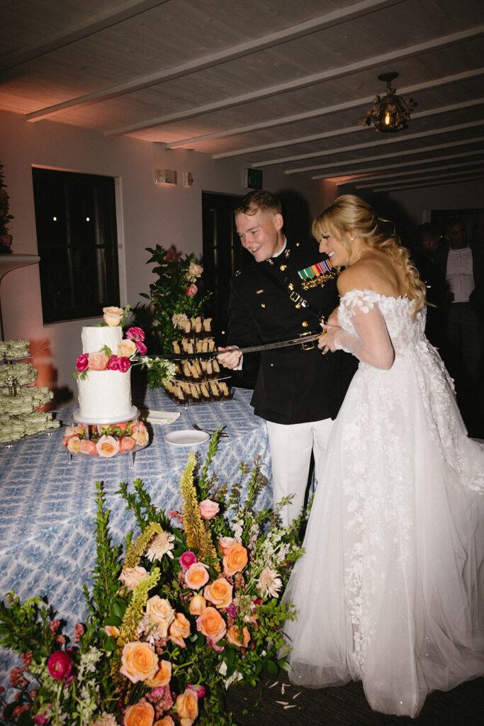 Bride and groom cutting cake together on dessert table with military sword.