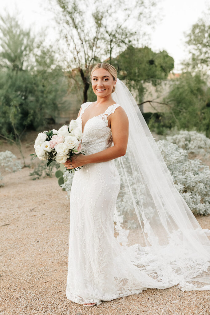 Bride standing in desert landscape, smiling, holding bouquet of white and blush flowers.