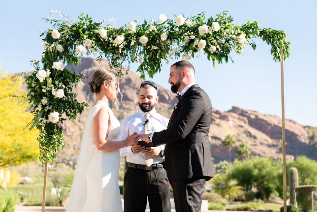 Bride and groom holding hands at outdoor wedding ceremony altar space with officiant behind them and arch with greenery and white flowers over them.