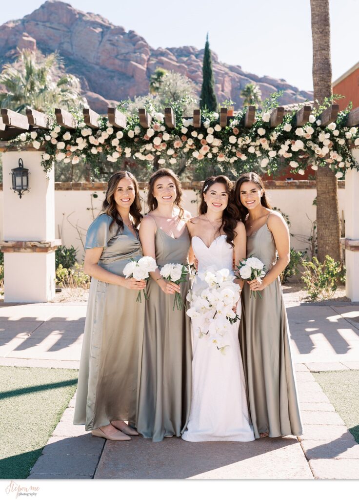 Bride with bridesmaids in sage green holding white flower bouquets with Camelback mountain in background at Omni resort.