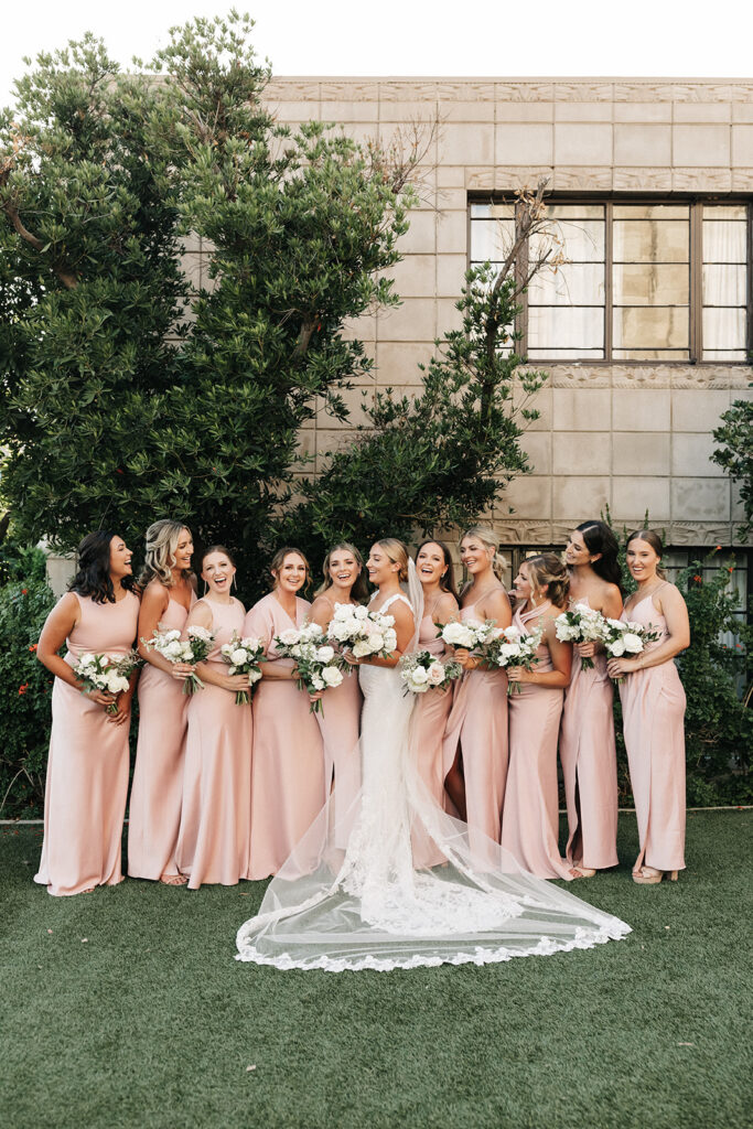 Bride standing in a line with bridesmaids, all holding bouquets of white and blush flowers, smiling.