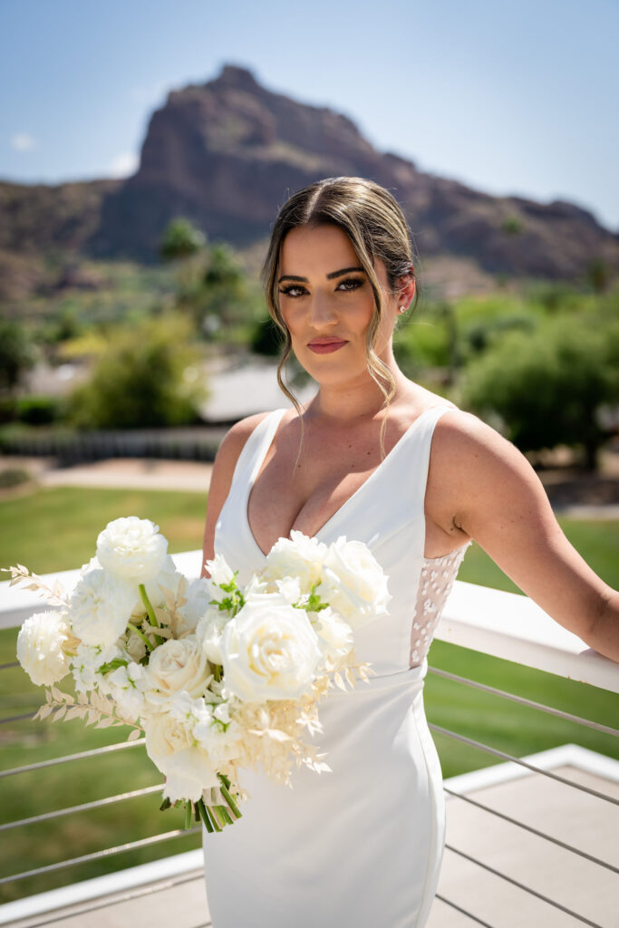 Bride standing in corner of patio railing at Mountain Shadows with Camelback Mountain in the background, holding white flowers bouquet.