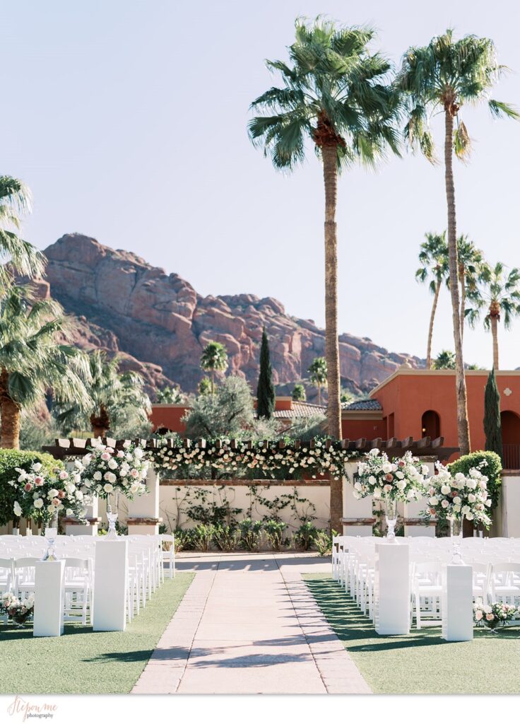 Outdoor wedding ceremony with two tall floral arrangements on either side of aisle in glass vases resting on white acrylic pillars.