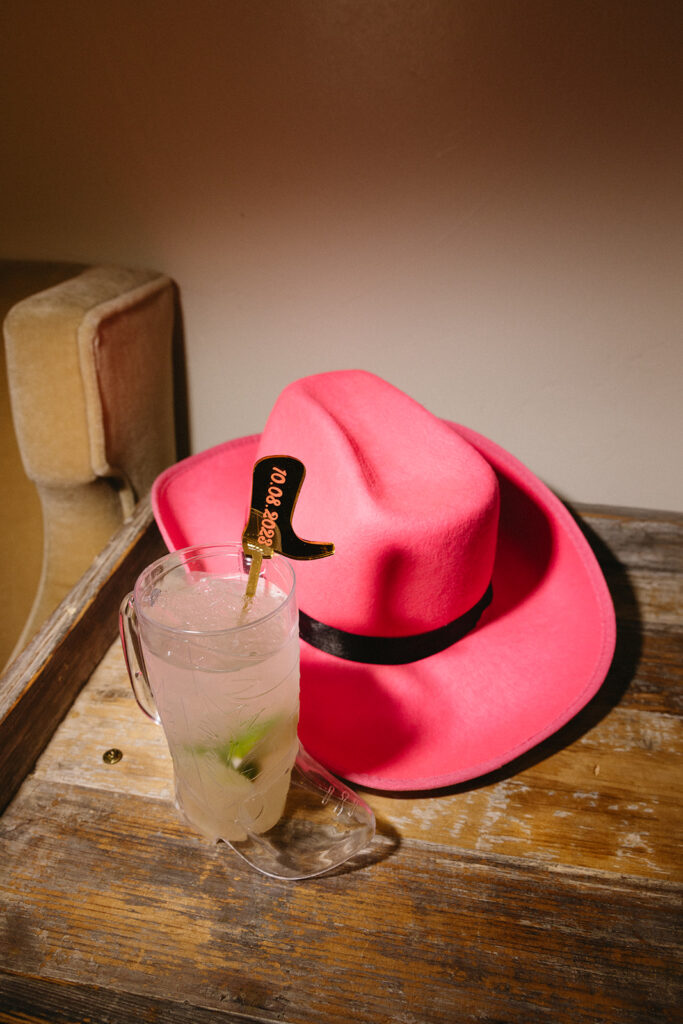 Pink cowboy hat on wood table with filled cowboy boot cocktail glass next to it.