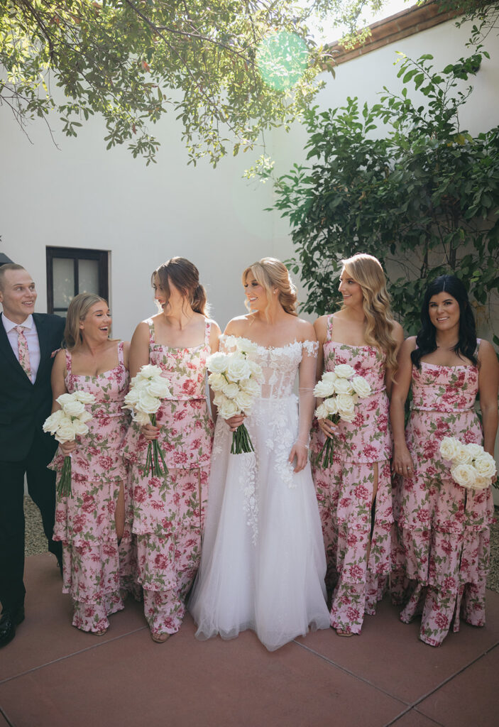 Bride walking with bridesmaids in pink floral dresses and bridesman on end with pink floral tie.