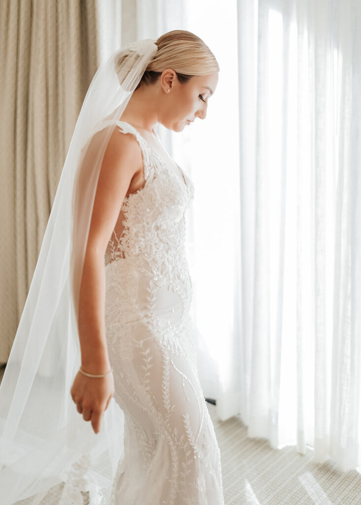 Bride standing in front of large windows with curtains drawn as light fills space, looking down in gown.