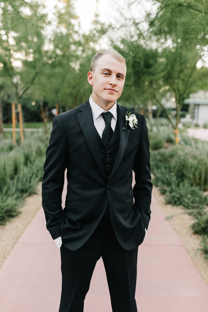 Groom in black suit with black bow tie and white flower boutonniere standing on paved path.