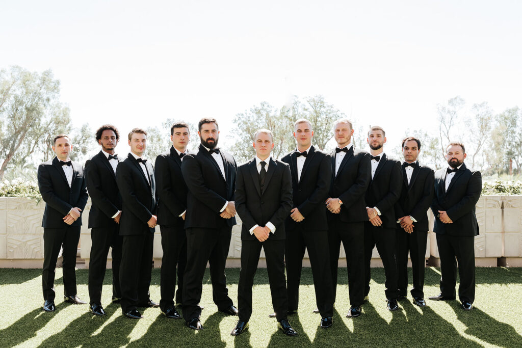Groom standing in center of line of groomsmen, all wearing black suits, groomsmen in black bow ties and groom in black tie.
