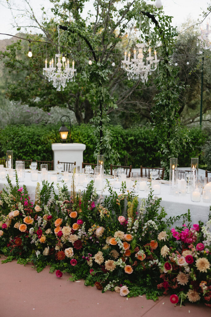 Ground floral placed in front of outdoor wedding reception table with white candles on table and chandeliers hanging over it.