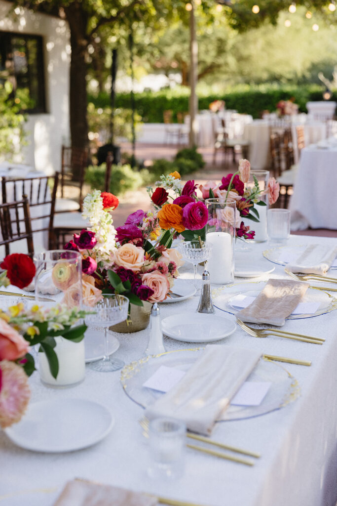 Colorful floral centerpieces in gold vases down center of wedding reception table with white linens.