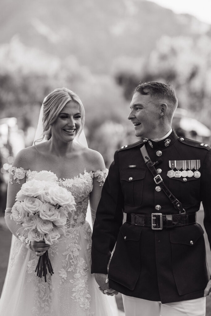 Bride and groom in military uniform holding hands, smiling, bride holding bouquet of roses with long stems.
