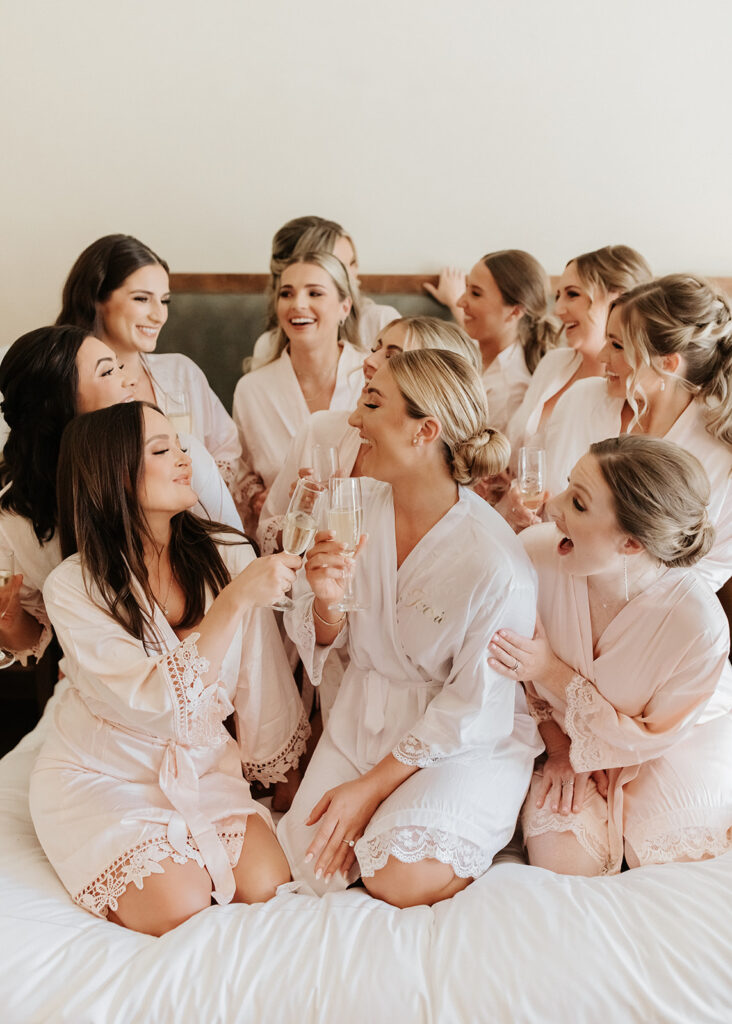 Bride in center of bridesmaids on a bed, all wearing bathrobes while getting ready for wedding.