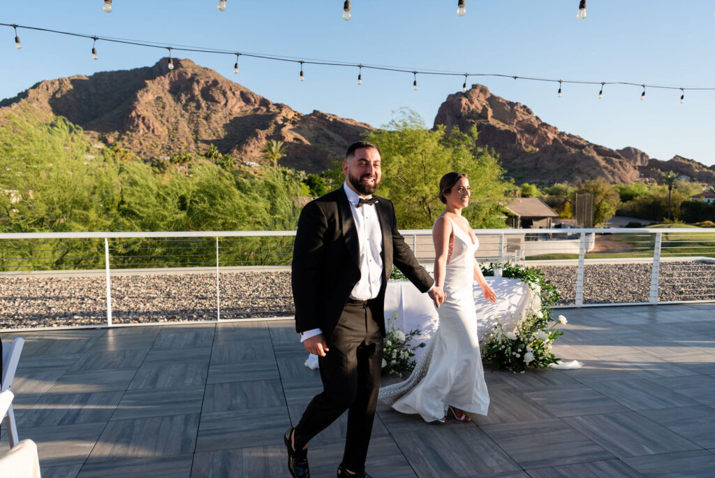 Bride and groom holding hands, walking into outdoor wedding reception patio with Camelback Mountain in the background.