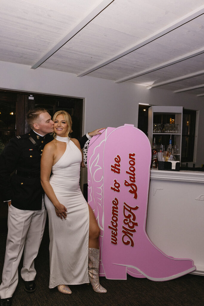Bride and groom standing next to large pink cowboy boot sign, groom kissing bride on cheek.