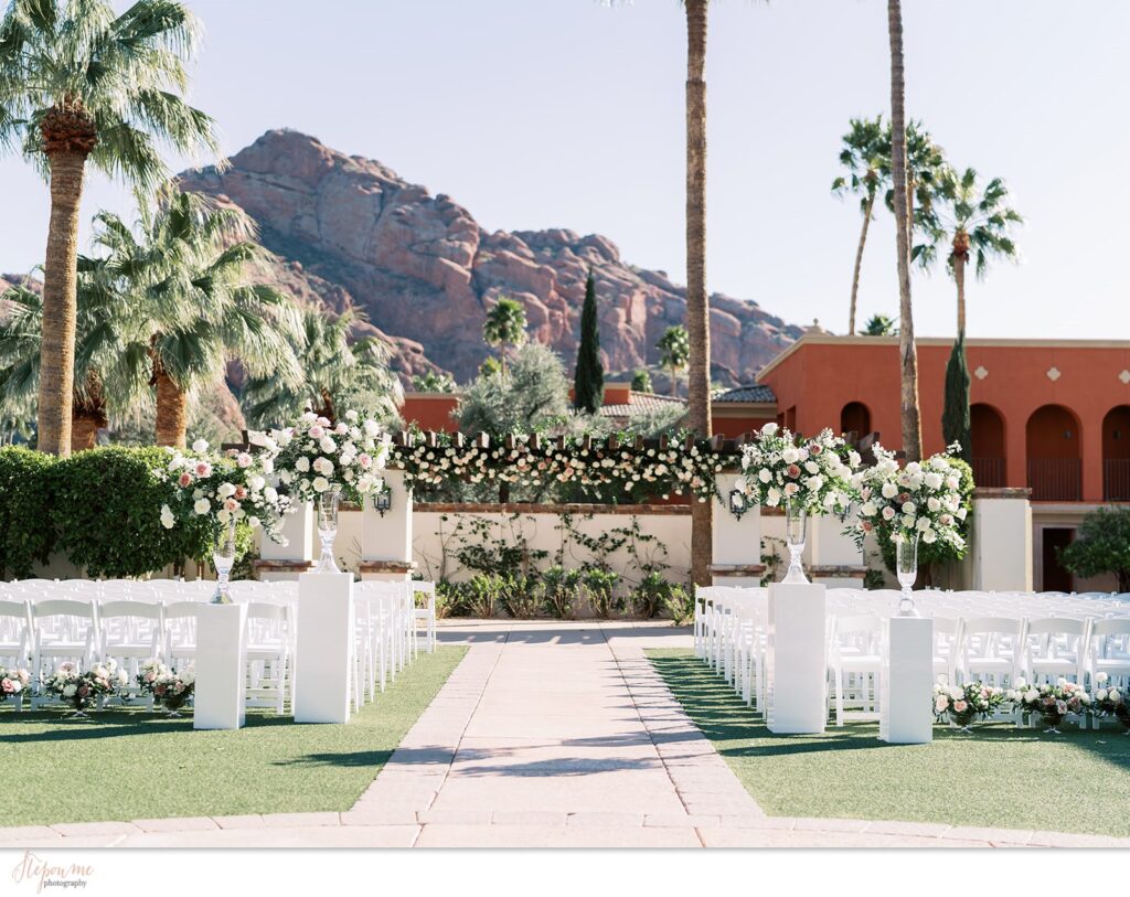 Outdoor wedding ceremony with two tall floral arrangements on either side of aisle in glass vases resting on white acrylic pillars.