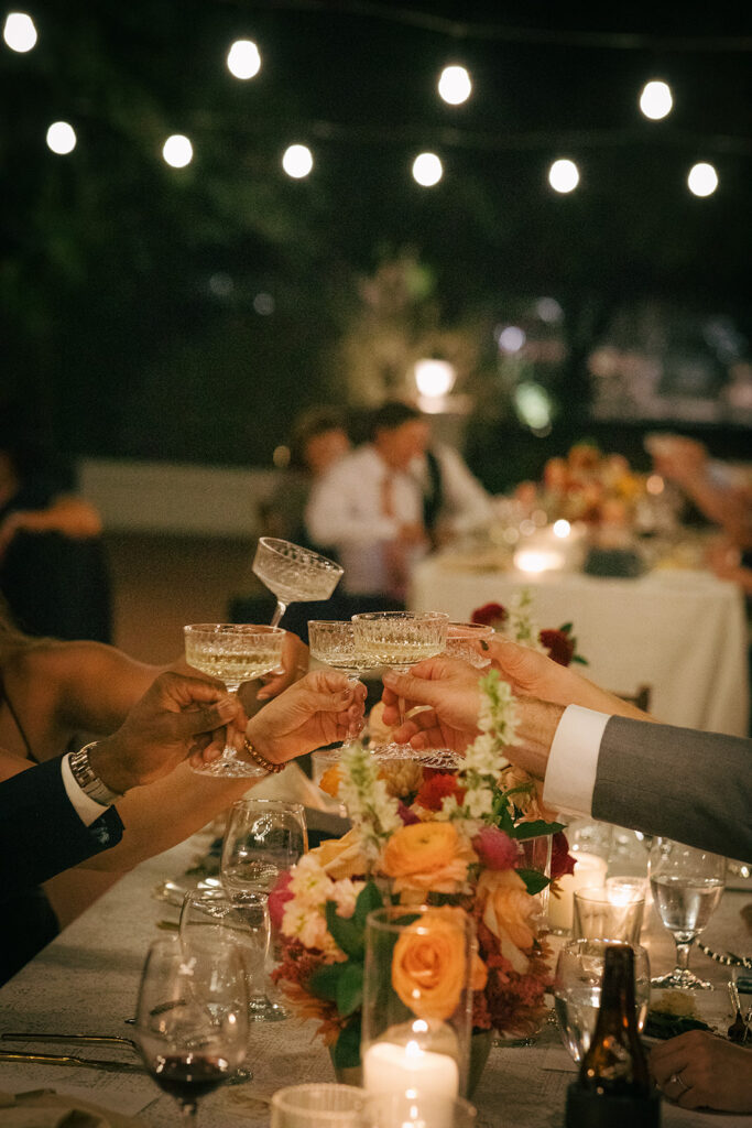 Wedding guests toasting glasses of champagne at outdoor reception table in the evening with hanging lights overhead.