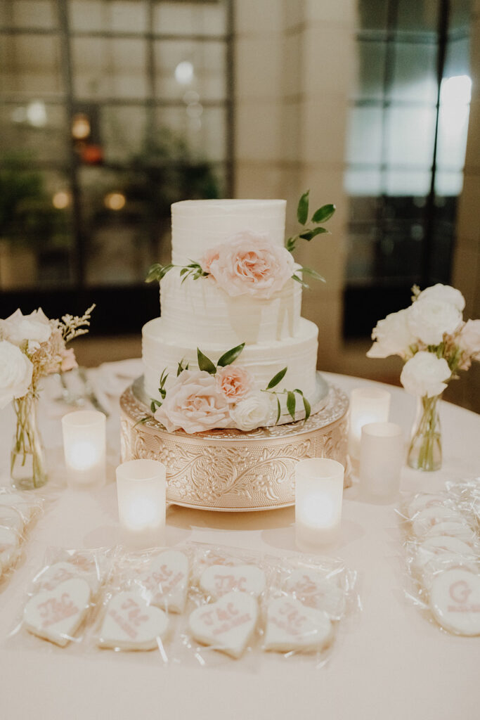 Three tiered white wedding cake with flowers added to it on table with custom cookies in bags on table in front of it, bud vases of flowers and votive candles around it.