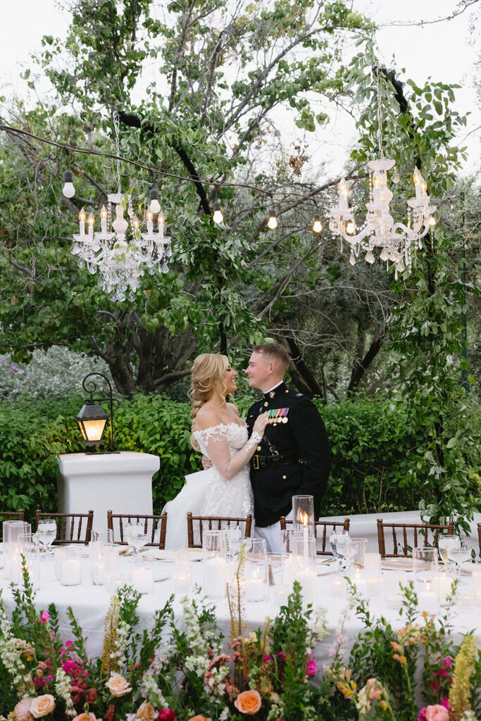 Bride and groom standing behind outdoor wedding reception table with white candles on table and chandeliers hanging over it and ground floral placed in front.