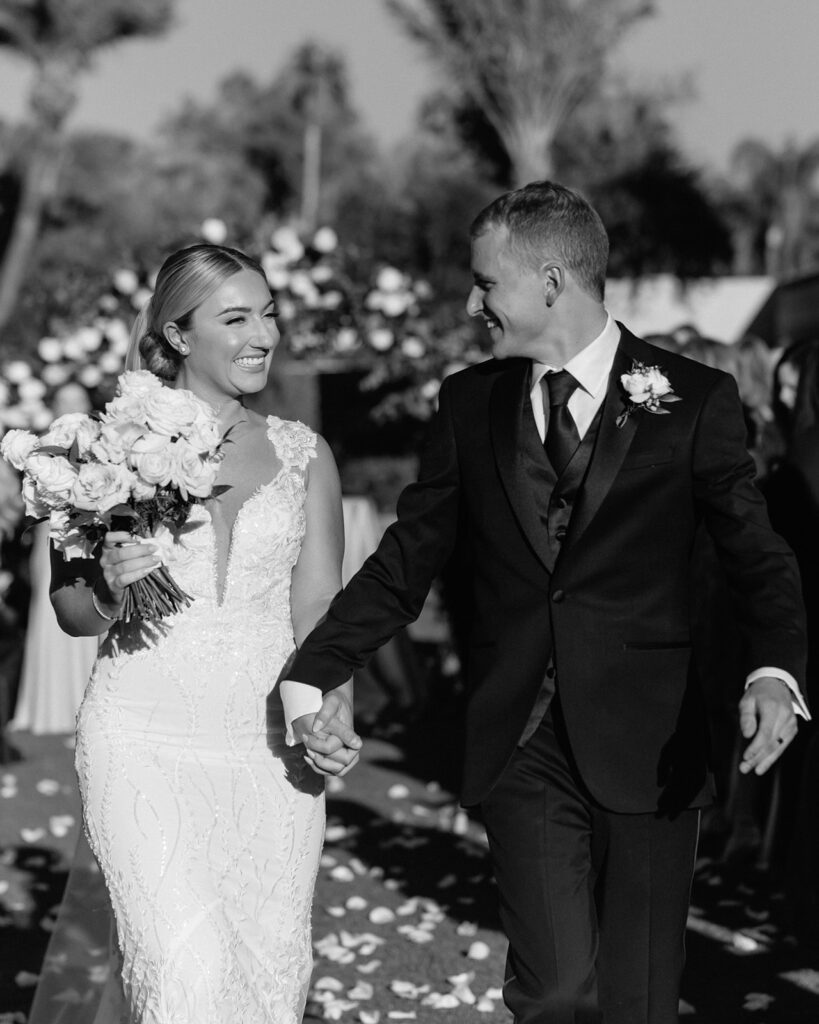 Bride and groom holding hands, smiling, while walking down wedding ceremony aisle.