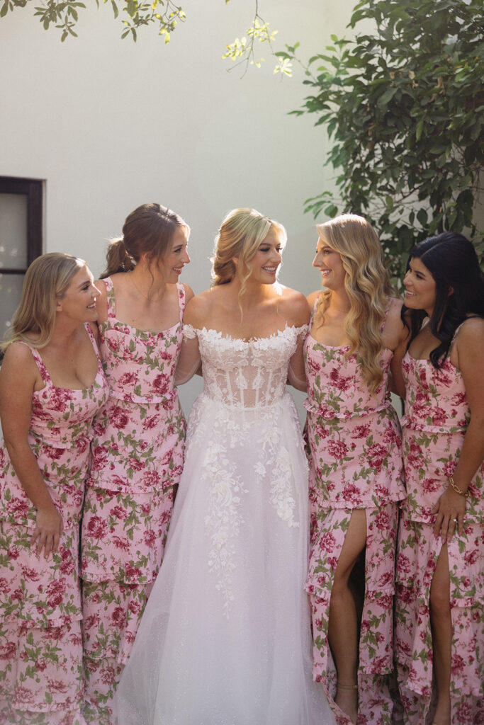 Bride with bridesmaids, all with arms around each other, bridesmaids wearing pink dresses with floral print.