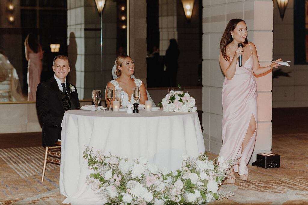 Bridesmaid giving speech next to sweetheart table with bride and groom sitting at it.