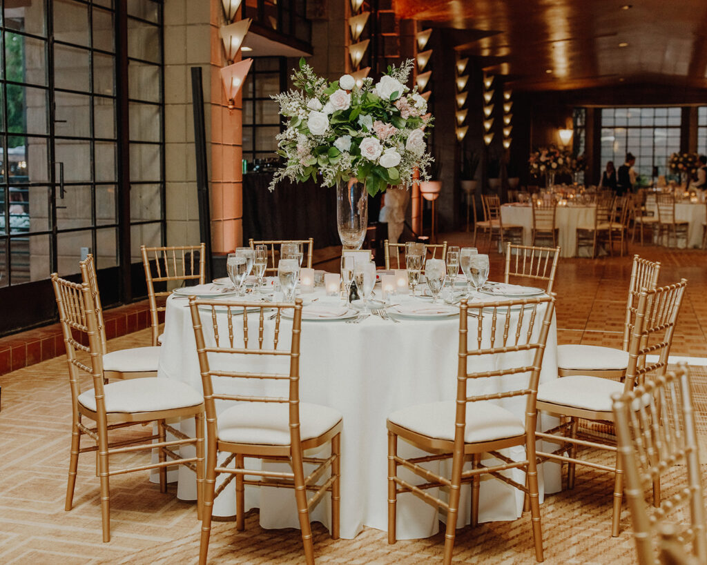 Indoor wedding reception space of white linen round tables with tall floral centerpiece on glass vase in center.