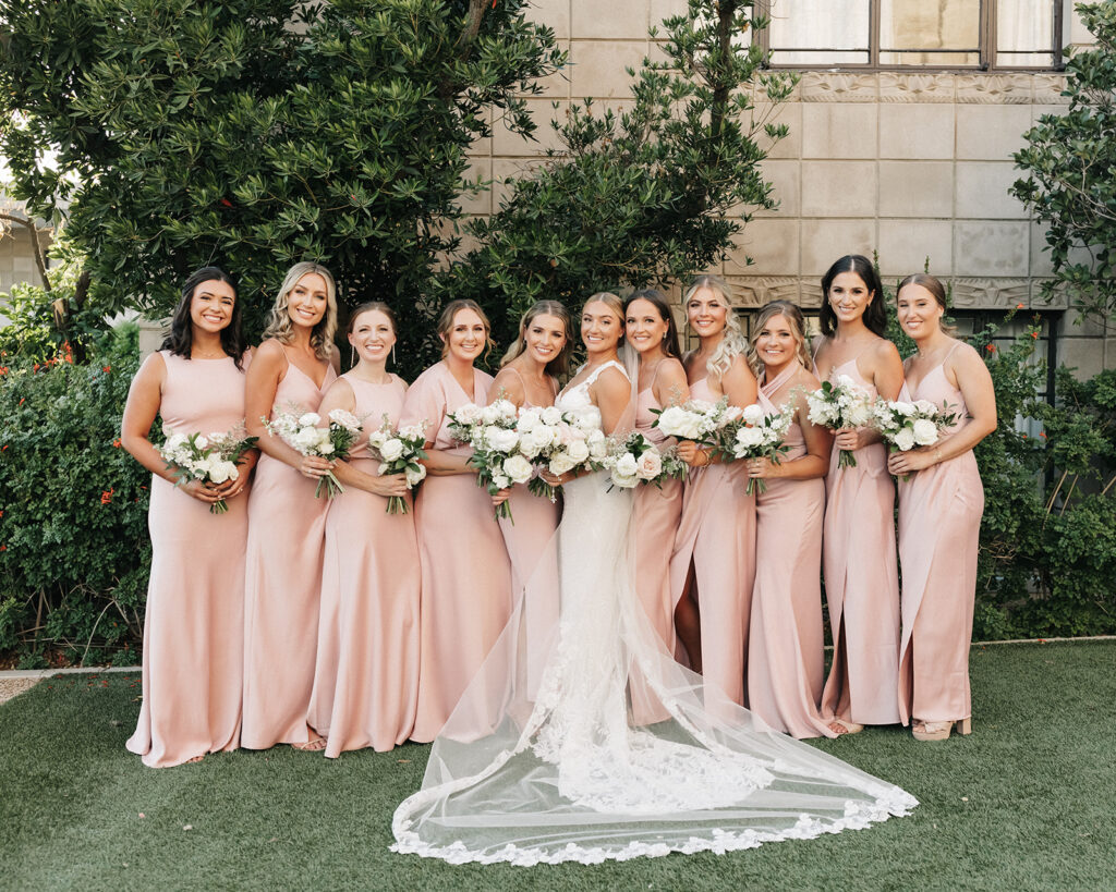 Bride standing in a line with bridesmaids, all holding bouquets of white and blush flowers, smiling.