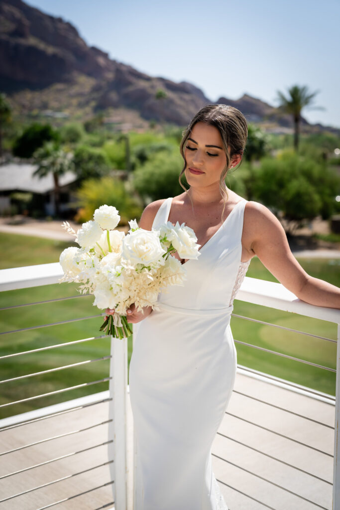 Bride standing in corner of patio railing at Mountain Shadows with Camelback Mountain in the background, holding white flowers bouquet.
