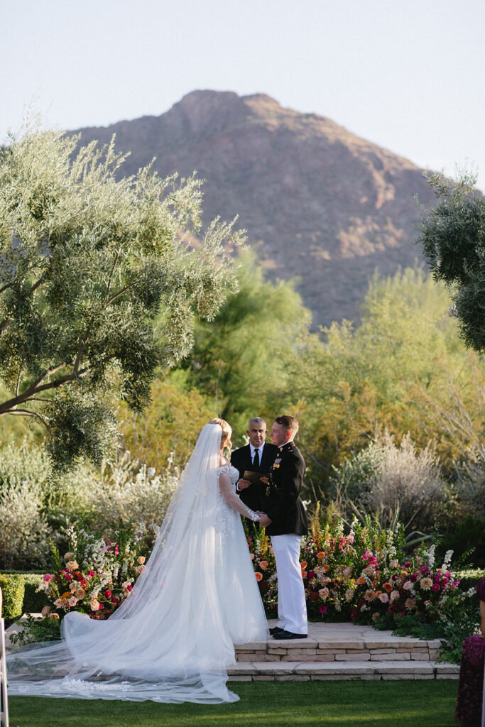 Bride and groom holding hands, standing at outdoor wedding ceremony altar space at El Chorro with officiant and Camelback Mountain in the background.