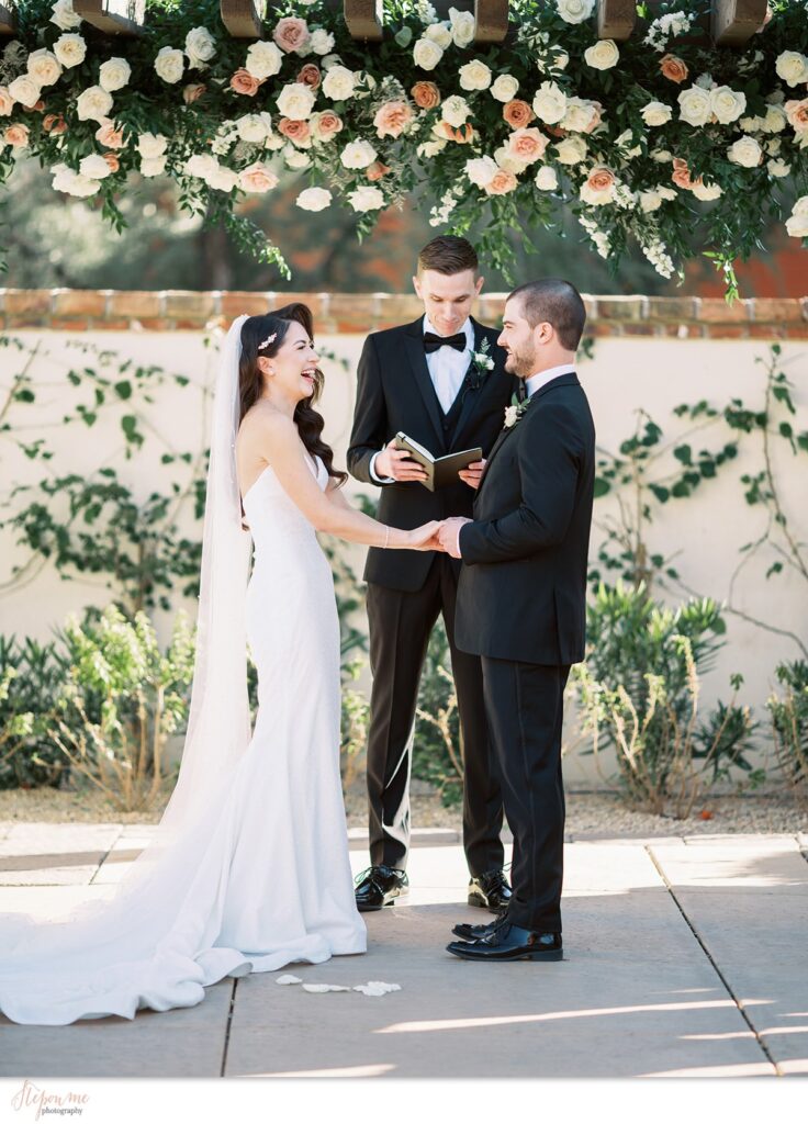 Bride and groom holding hands under pergola with flowers of white and pink, mauve installed on it.