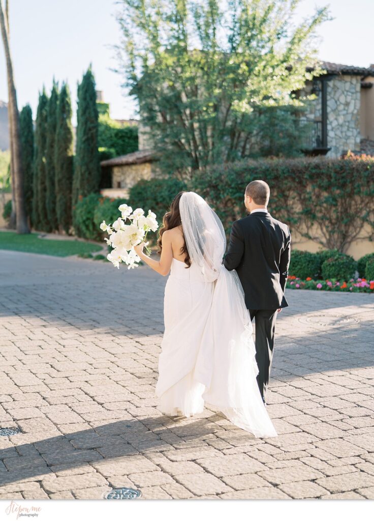 Bride and groom walking away holding hands, bride holding bouquet, on paved brick space.