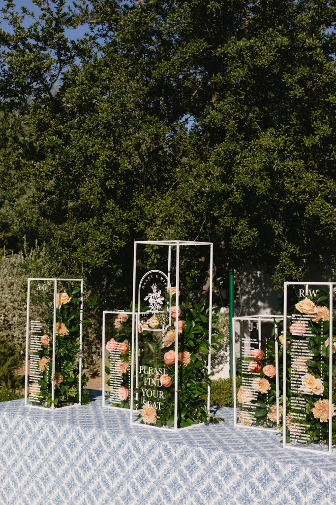 Custom wedding escort sign clear columns with floral and greens in center and guest names listed on front.